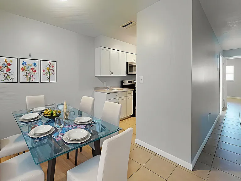 Modern dining area with a sleek glass table, elegant white chairs, and a cozy kitchen, perfect for stylish entertaining.