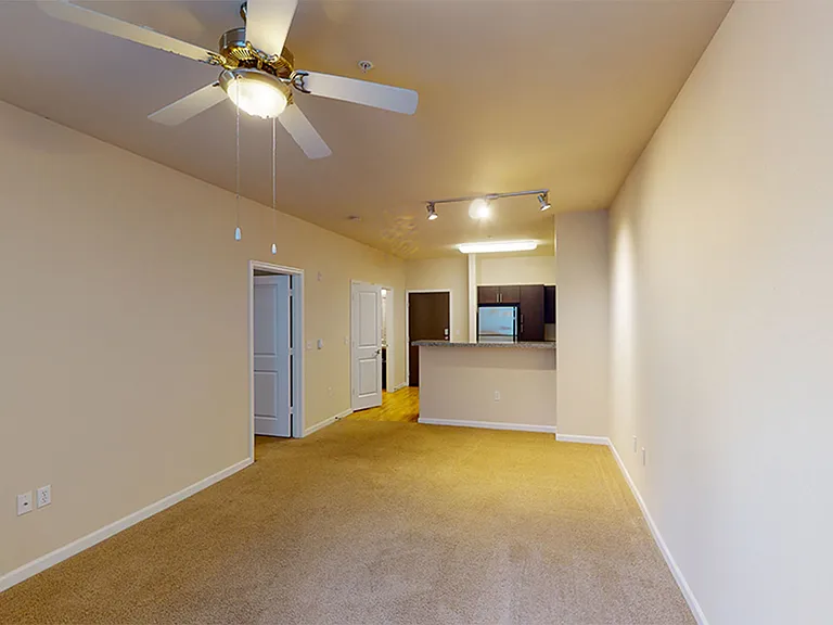 Apartment living area with carpet looking into kitchen with hardwood and dark cabinets.