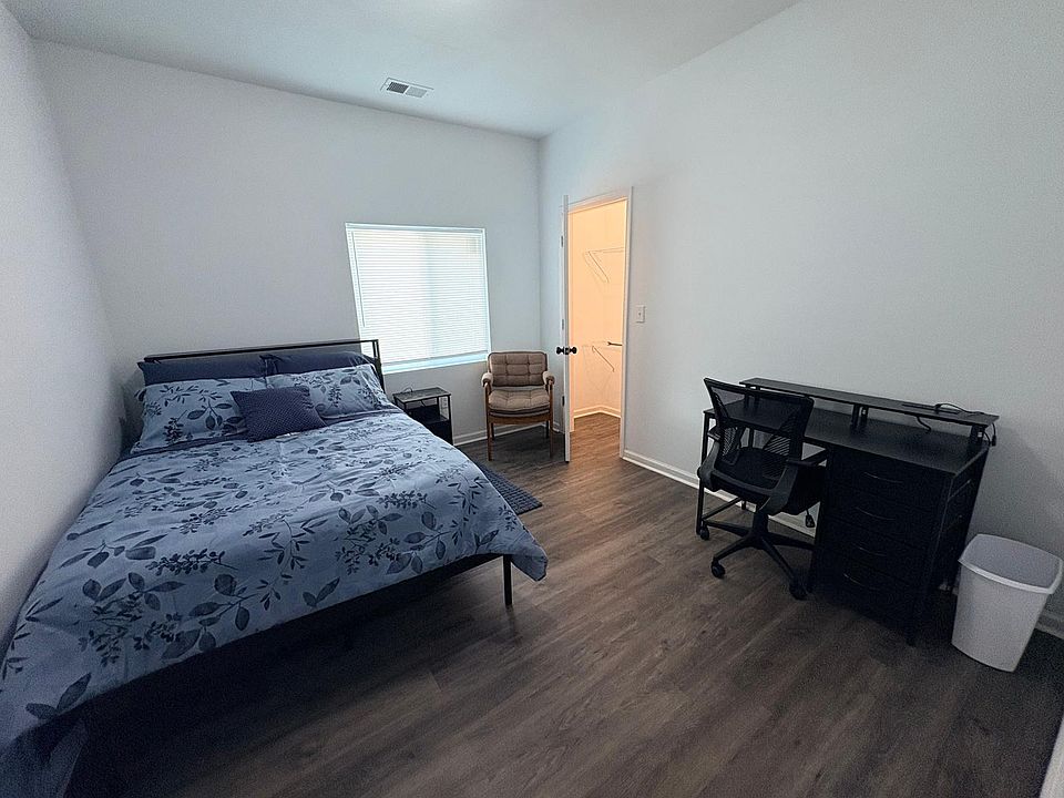 Furnished bedroom with full bed, work desk, and a walk-in closet, plus a window with blinds and wood-look flooring.