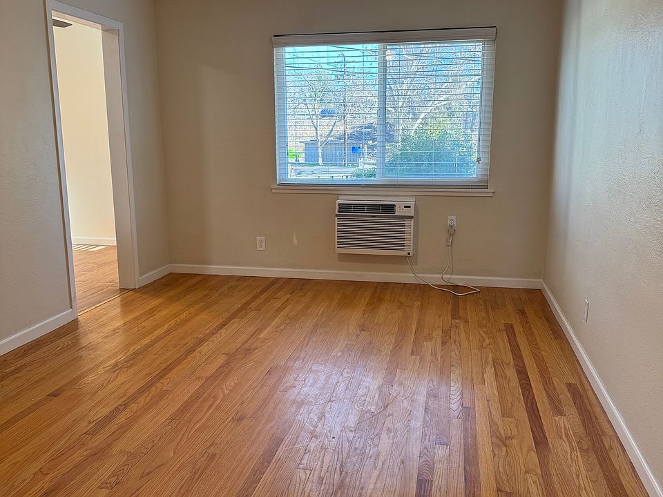 Looking at living room window and wall AC unit, new ceiling fan, from front door. Doorway to kitchen and nook on left. Original hardwood floor.