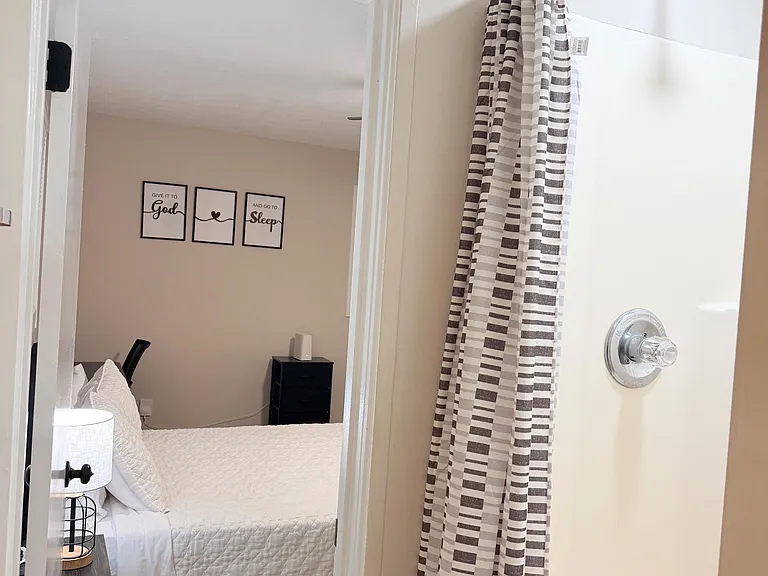 Bathroom with a tub/shower combo and striped shower curtain, located near a furnished bedroom with bedside lighting.