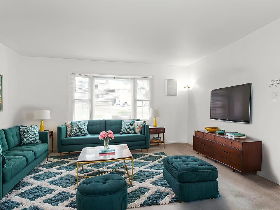 Bright and inviting living room featuring teal sofas and ottomans, a stylish coffee table with pink flowers, and large windows for ample natural light.