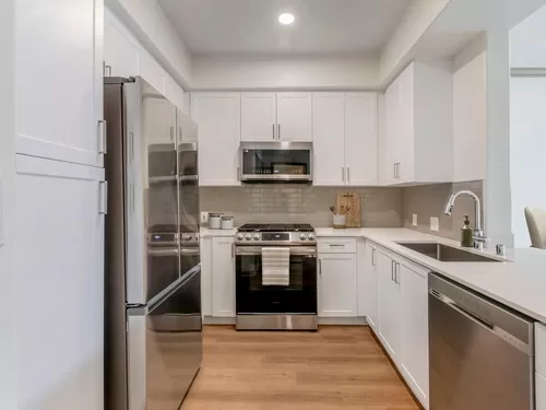 Renovated Package I kitchen with stainless steel appliances, white speckled quartz countertop, white cabinetry, grey tile backsplash, and hard surface flooring - Avalon Thousand Oaks Plaza