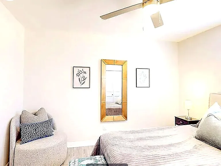 Warm, inviting private bedroom with accent chair, gold-framed mirror, ceiling fan, and tasteful decor