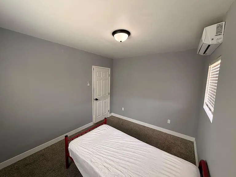 Private bedroom with carpeted floors, gray walls, window blinds, and a wall-mounted mini-split for efficient cooling.
