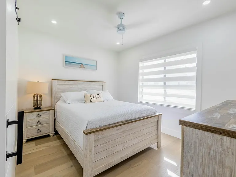 Private bedroom with a full bed, nightstand, and dresser, plus a large window with layered blinds and ceiling fan.