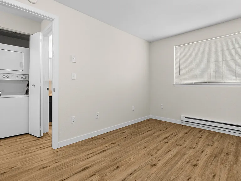 Bright bedroom with wood-style plank flooring, baseboard heating, window with blinds, and direct access to in-unit stacked washer and dryer at Garden Square Apartments in Lakewood, WA. Bright bedroom with wood-style plank flooring, baseboard heating, wind