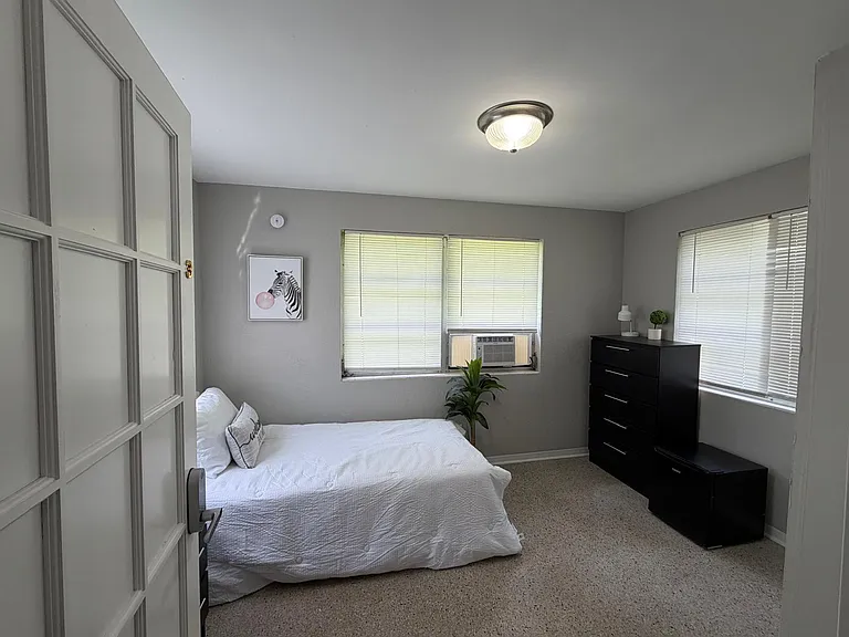 Private bedroom with a full bed, dresser, and mini fridge, plus dual windows and an in-room window A/C unit.