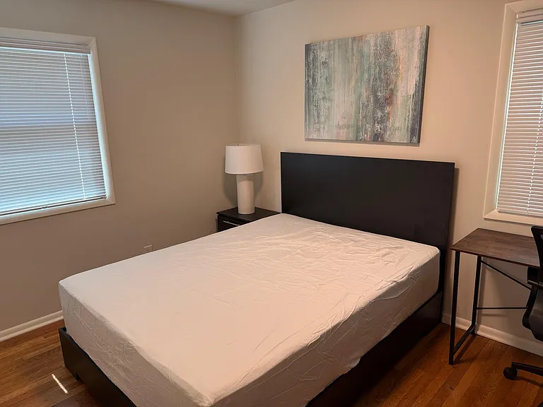 Furnished bedroom with a full bed, nightstand, and desk, plus two windows with blinds for natural light and privacy.