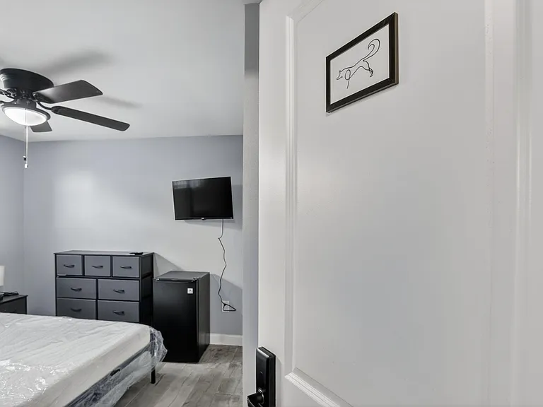 Bedroom includes a ceiling fan, dresser storage, wall-mounted TV, and a mini fridge for added in-room convenience.