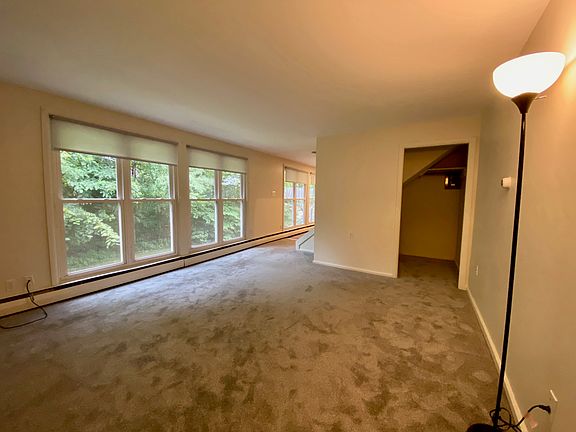 Large living room with new grey wall to wall carpet, closet area and lots of windows with shades.
