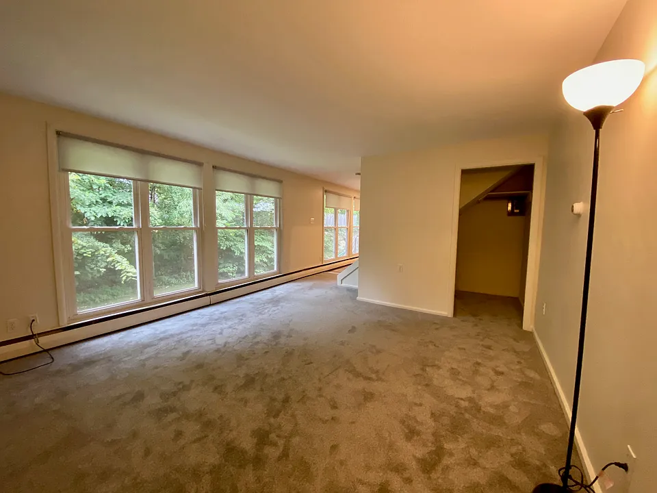 Large living room with newer grey wall to wall carpet, closet area and lots of windows with shades.