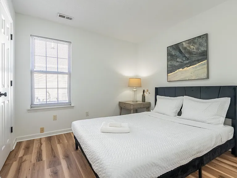 Private bedroom with a furnished bed, nightstand and lamp, plus a window with blinds and durable wood-look flooring.