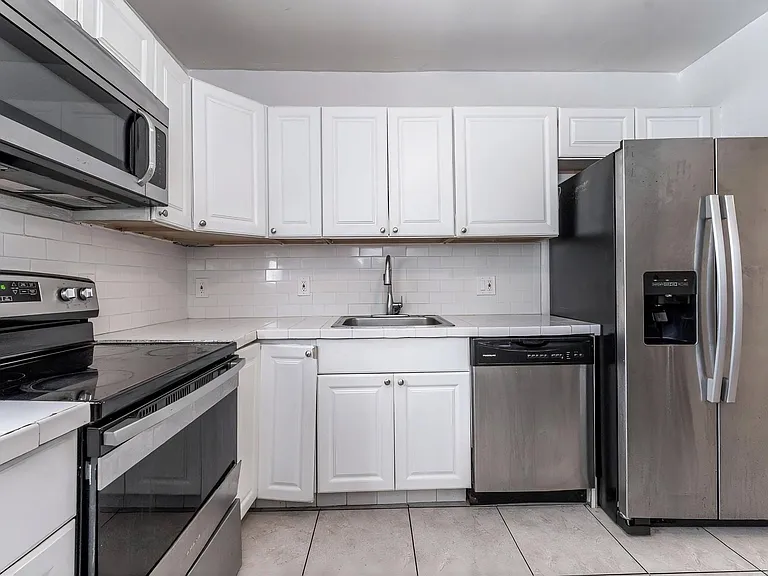Efficient galley kitchen with ample white cabinetry, tiled counters and backsplash, and a full stainless steel appliance suit