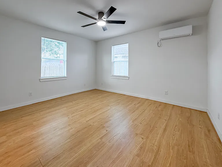 This room features light wood flooring, large windows for natural light, a modern ceiling fan, and a wall-mounted air conditioning unit, creating a bright and comfortable space.