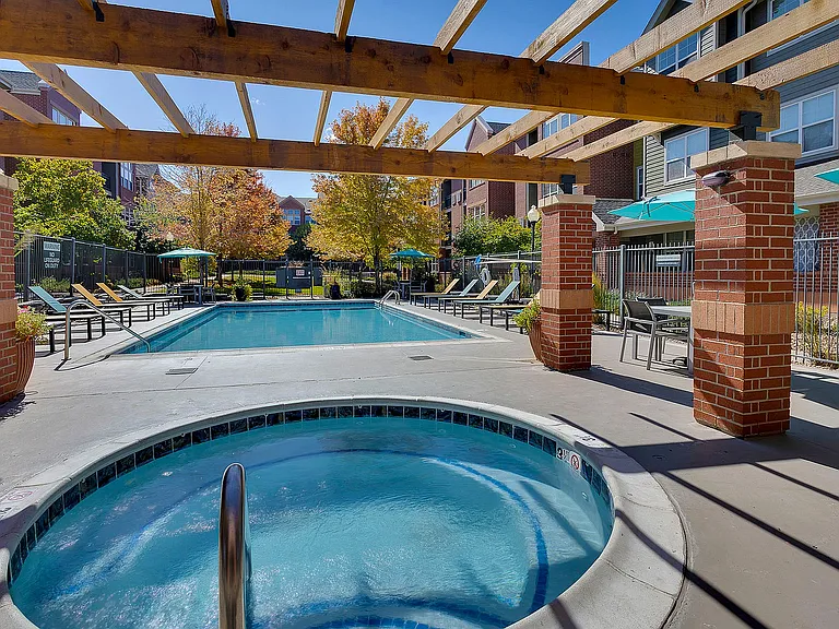Chic apartment complex featuring a sunlit outdoor pool area with relaxing lounge chairs and a cozy hot tub under a stylish pergola.