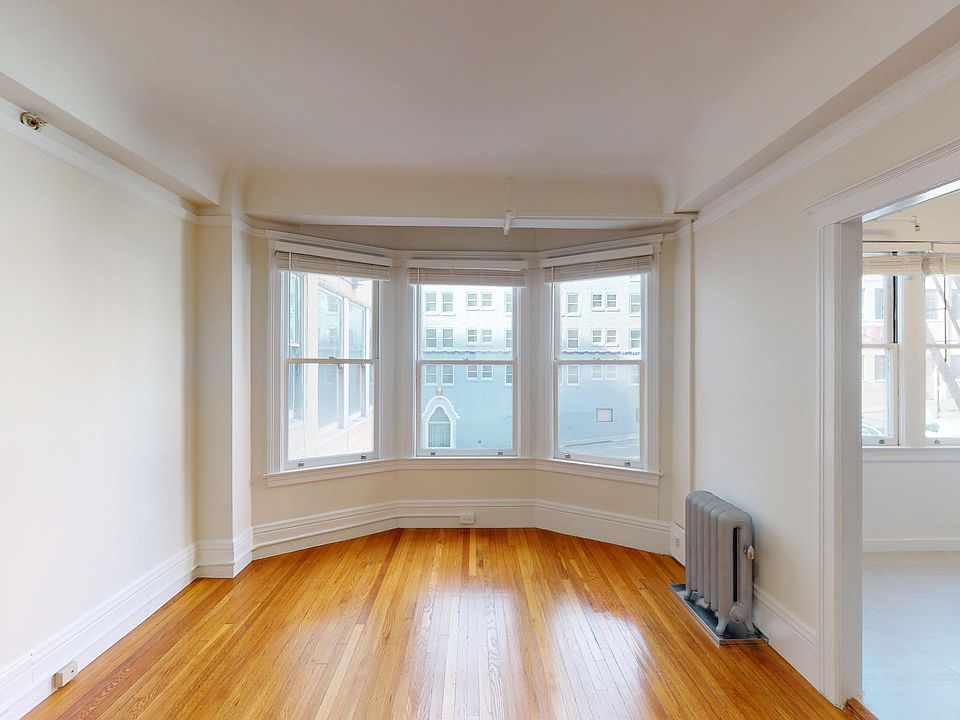 Bright living space with bay windows, hardwood floors, and a glimpse of a kitchen area. Elegant, minimalistic design.