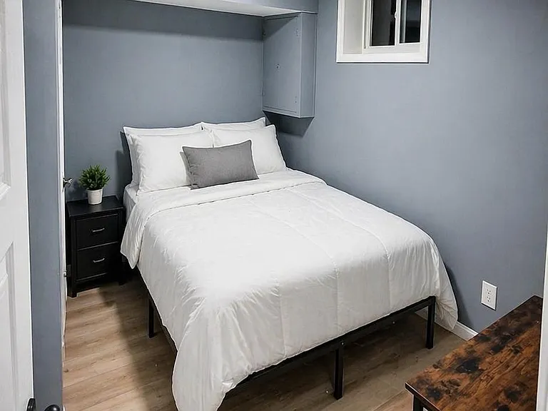 Compact bedroom with a queen bed, bedside table, and desk, plus a small window and wood-look flooring.