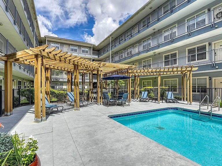 Charming apartment complex courtyard featuring a pristine swimming pool, wooden pergola, and relaxing lounge chairs under a bright blue sky.