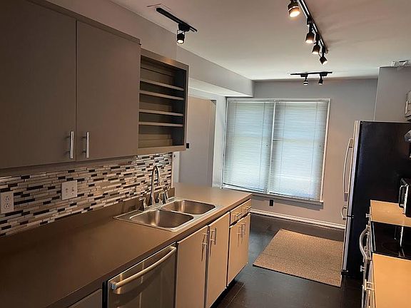 View looking back through kitchen from living room entrance, track LED lighting, updated sink fixture, Bosch dishwasher, accent backsplash, and window mini blinds.