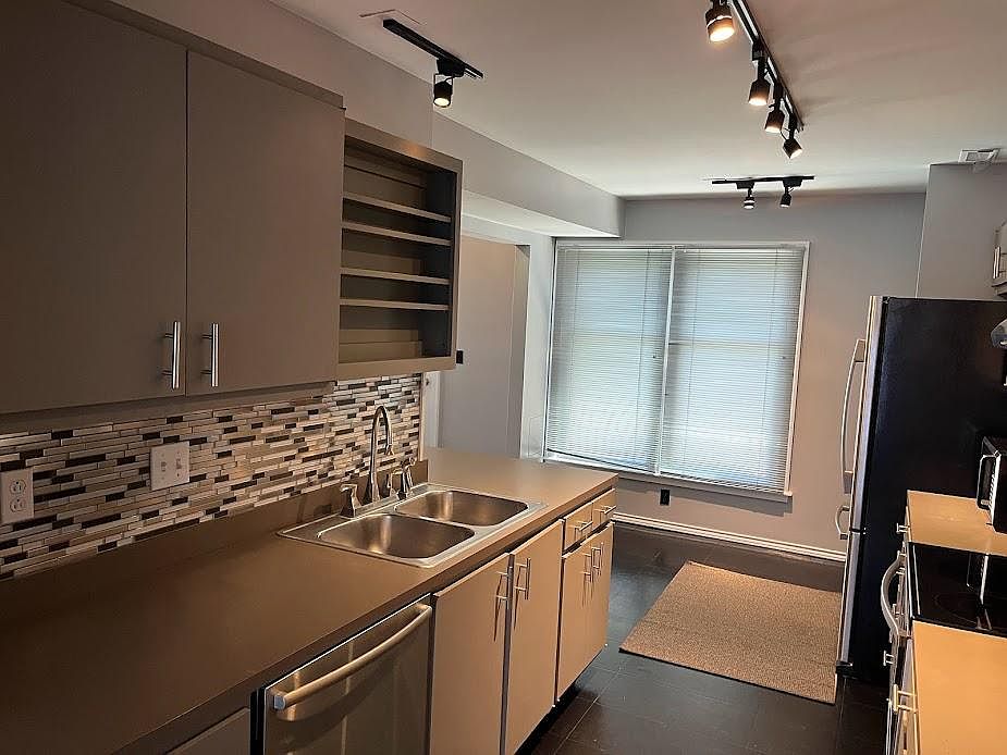 View looking back through kitchen from living room entrance, track LED lighting, updated sink fixture, Bosch dishwasher, accent backsplash, and window mini blinds.