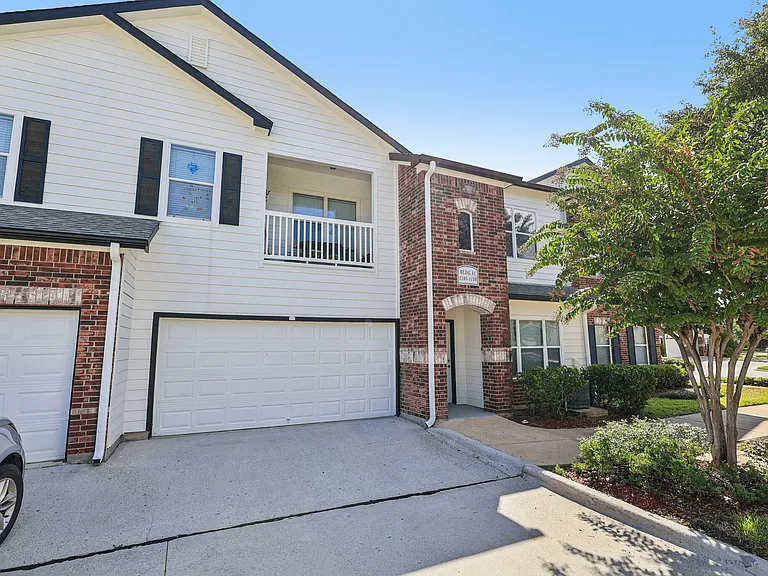 Charming townhouse exterior showcasing a blend of white siding and red brick with lush greenery and a two-car garage. Charming townhouse exterior showcasing a blend of white siding and red brick with lush greenery and a two-car garage.