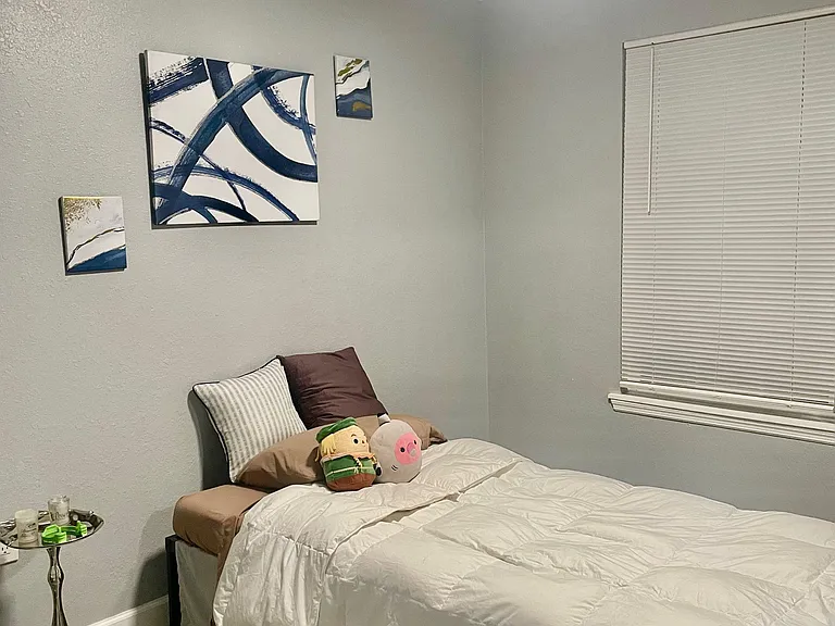 Furnished bedroom with a comfortable bed, nightstand, wall art, and a window with blinds for light control.
