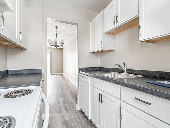 Modern kitchen with electric stove, dishwasher, refrigerator, faux granite countertops, white cabinetry, and view to dining/living areas
