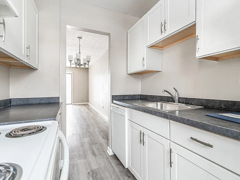 Modern kitchen with electric stove, dishwasher, refrigerator, faux granite countertops, white cabinetry, and view to dining/living areas