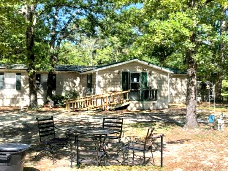Single-story home set on a shaded lot with a ramped entry and outdoor seating area for relaxing or gathering.