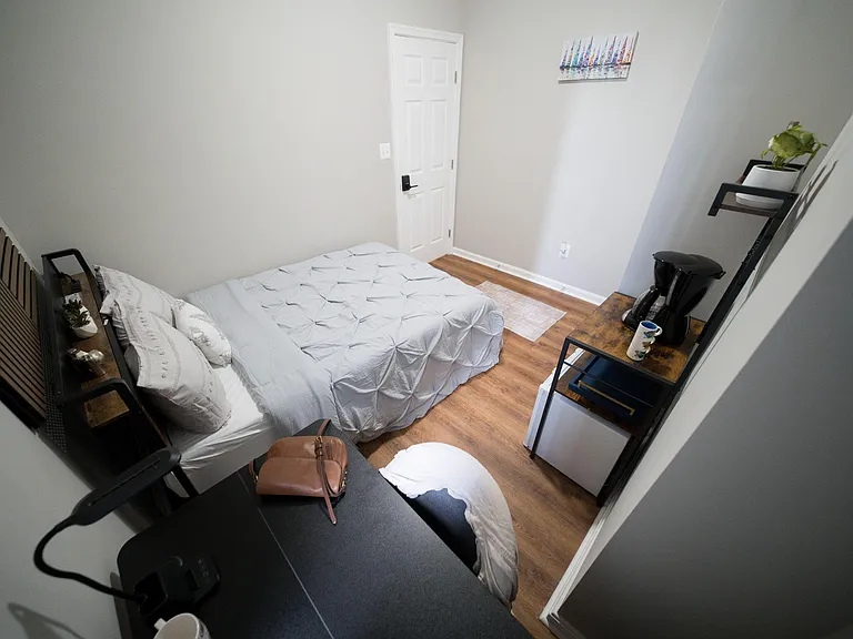 Furnished bedroom with a full bed, hardwood-style floors, and a compact workstation plus mini-fridge and coffee station.
