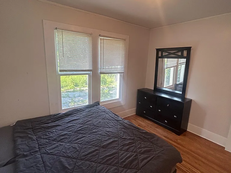 Furnished bedroom with two large windows, hardwood floors, and a dresser with mirror for convenient storage.