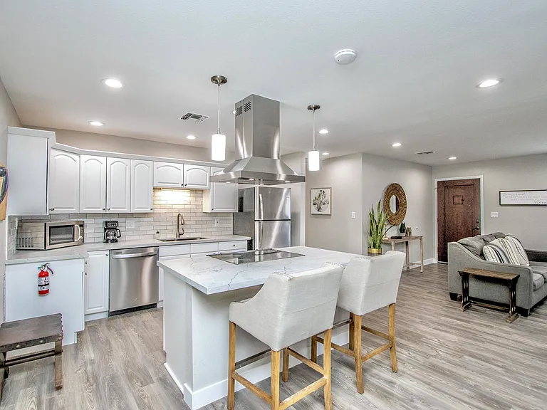 Sleek kitchen view at Casa Del Sol, Tempe rental home. Showcases a quartz island with breakfast bar stools, stainless steel appliances, and white cabinetry, flowing seamlessly into the open-concept living area. Perfect for a premium Tempe, AZ stay.