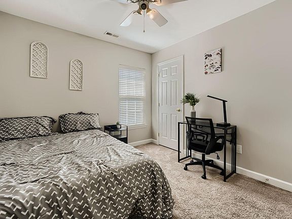 Carpeted bedroom with a comfortable bed, window with blinds, and a dedicated desk setup for work or study.