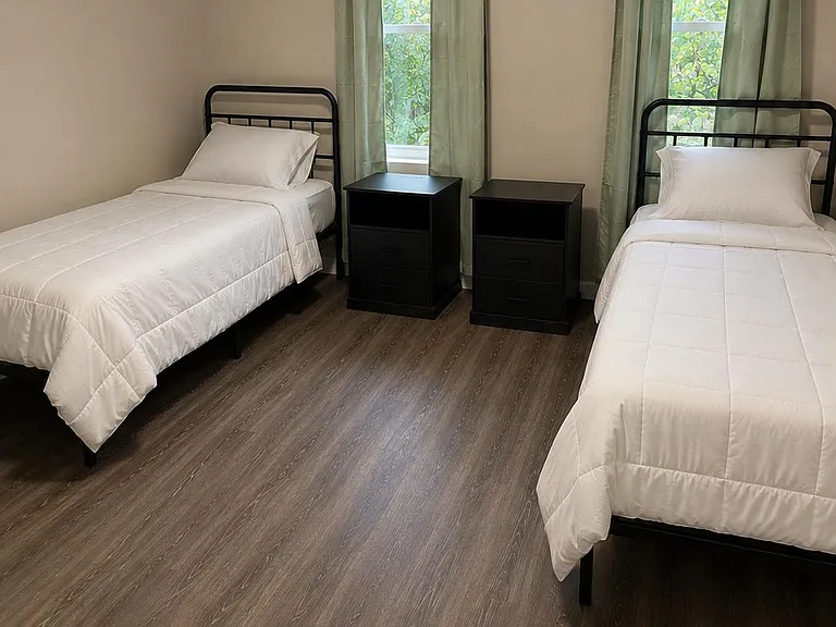 Spacious shared bedroom with two twin beds, wood-style flooring, and two windows with curtains plus bedside storage.