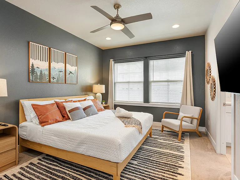 Bright and inviting bedroom with a king-sized wooden bed, white and burnt orange bedding, and matching pillows. Features two bedside lamps, a cozy armchair, a ceiling fan, a large window for natural light, and a wall-mounted TV for your comfort.