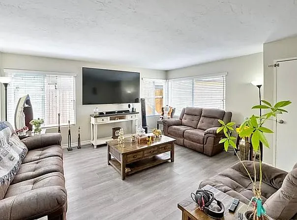 Spacious living room with multiple seating areas, large wall-mounted TV, and ample windows with blinds for natural light.