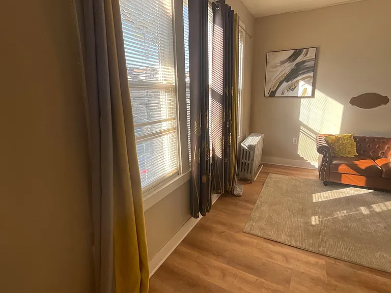 Living area with tall windows, curtain panels and blinds, plus wood-style flooring and space for comfortable seating.