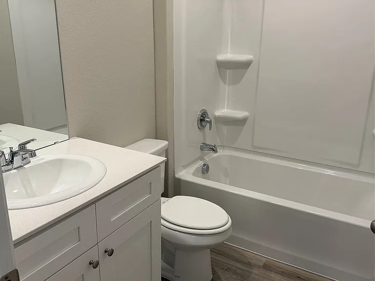 Bathroom featuring a white vanity with panel cabinetry and a contrasting countertop, a white toilet, a white bathtub with an integrated shower, chrome fixtures, and wood-finish flooring