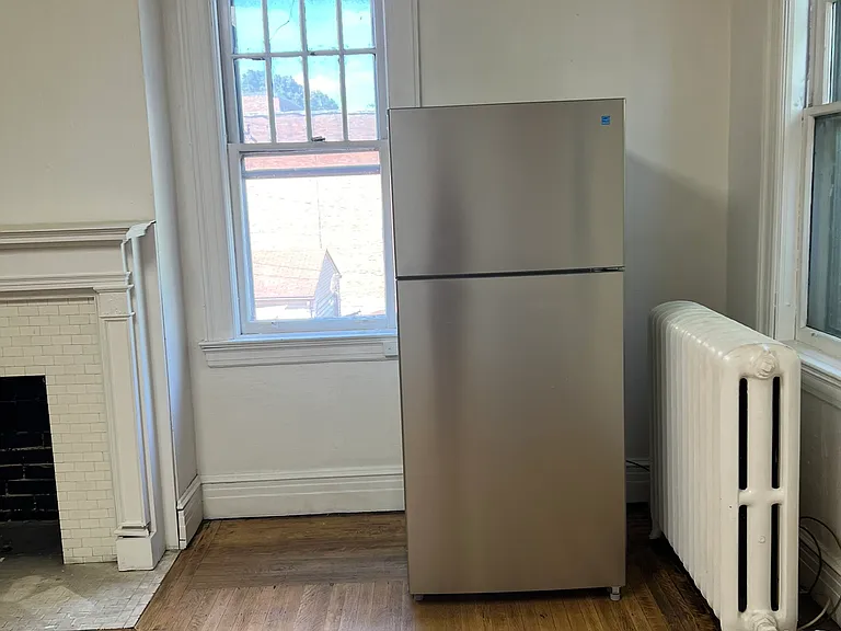Stainless-steel refrigerator included, positioned beside tall windows, classic radiator heat, hardwood flooring, and a decorative fireplace.