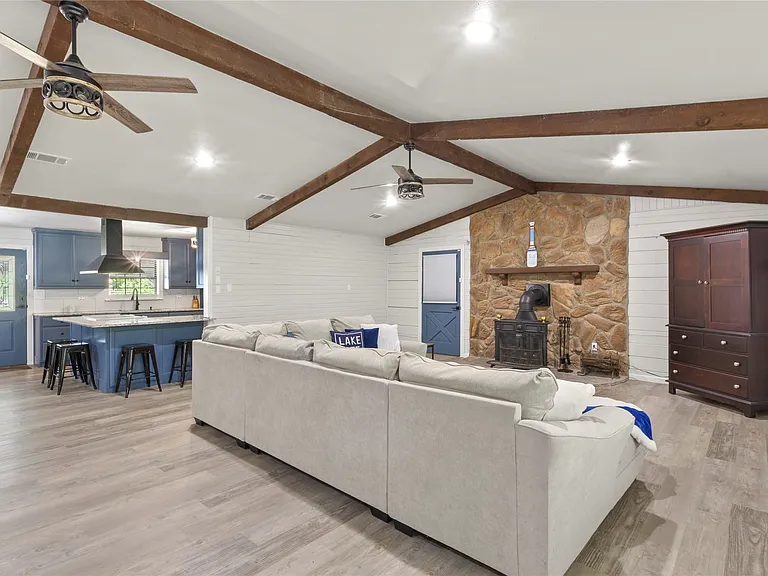 Spacious open-concept living area featuring a stone fireplace, exposed wood beams, and wood-finish flooring