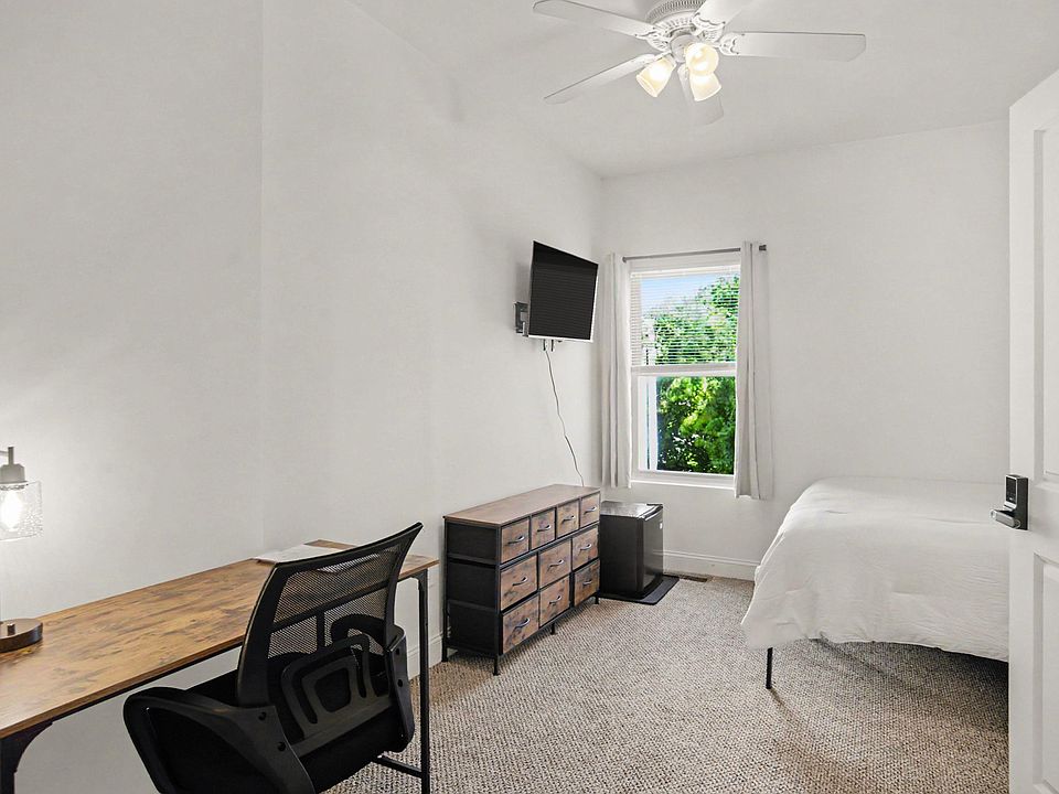 Furnished bedroom with a work desk, dresser storage, wall-mounted TV, and a window with blinds and curtains.