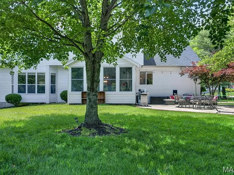 Spacious backyard featuring a large tree, vibrant green lawn, white siding, and a stone patio