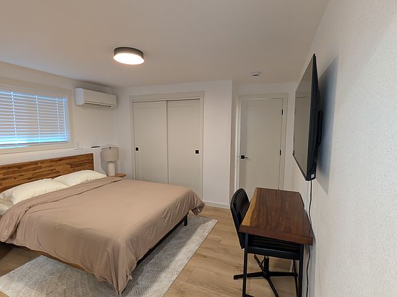 Shows the queen size bed, dedicated workspace, smart TV, wall mounted mini split, closet, side table with lamp.