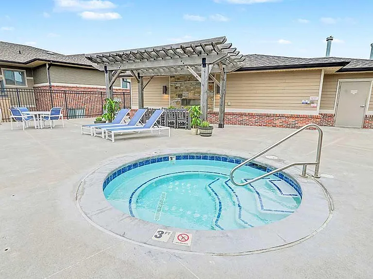 Apartment community pool area with adjacent circular hot tub, spacious concrete deck, and rows of blue lounge chairs enclosed by residential buildings and fencing.