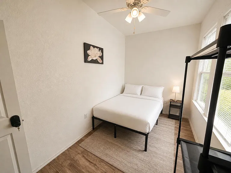 Private bedroom with a full bed, nightstand and lamp, plus a ceiling fan and large window with blinds for natural light.