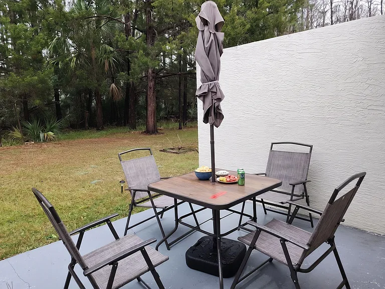 Covered patio with outdoor dining set and umbrella, opening to a grassy yard bordered by mature trees for privacy.
