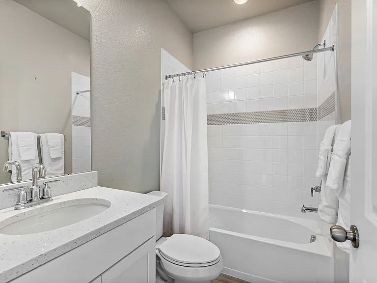 Clean and bright bathroom with white vanity, quartz countertop, and chrome fixtures. Features a bathtub with white tiled walls and decorative accent strip, a shower curtain, and neatly folded towels, creating a fresh and inviting atmosphere.