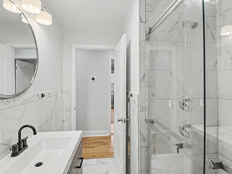 Updated bath with a glass-enclosed tub/shower, marble-look tile surround, and a wide vanity with modern lighting.