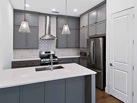 Kitchen featuring an island with an undermount sink, quartz countertops, gray shaker-style cabinetry, stainless steel appliances, and a subway tile backsplash